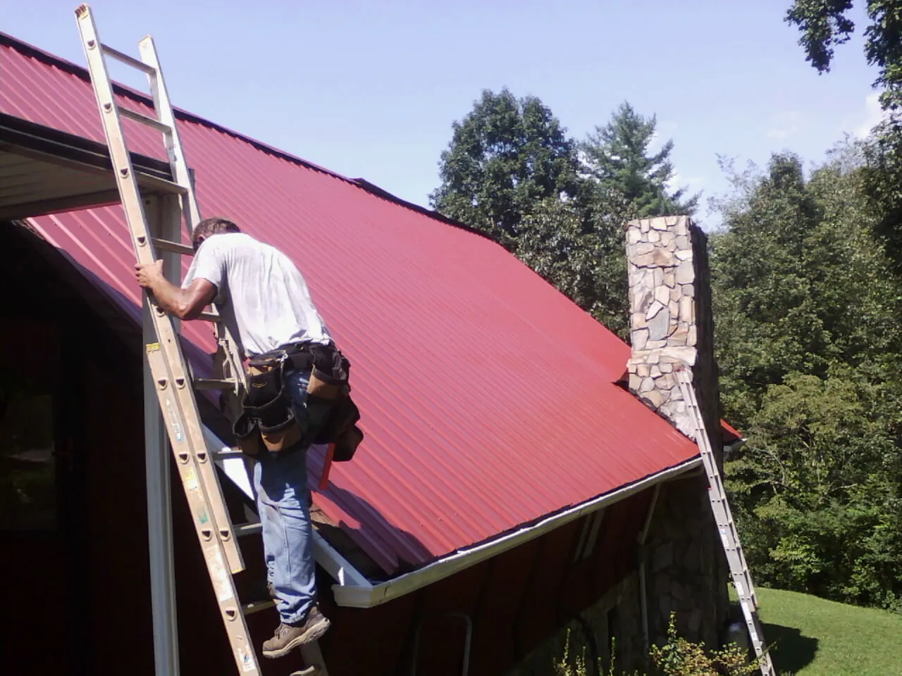 Roofer on a ladder beside a red metal roof and stone chimney — Soffit & Fascia Repair by Roofing Brothers in Gustine